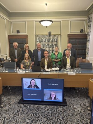 A group of people standing and sitting behind a desk, with two more joining the session remotely on a screen